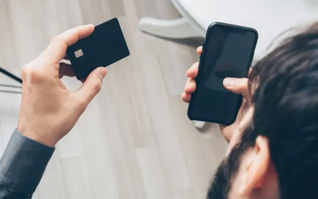 O persoană ține în mâna stângă un card bancar negru cu cip, iar în mâna dreaptă un telefon mobil cu ecranul stins sau negru